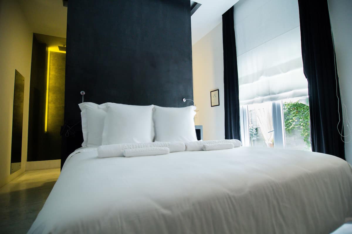 A neatly made bed with white linens and pillows, a black headboard, and a view of greenery outside through a large window.