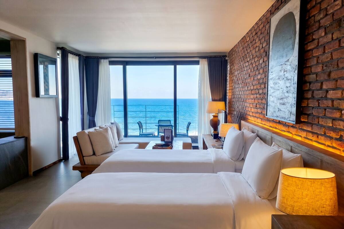 Une chambre d’hôtel avec deux lits, un balcon donnant sur la mer, un mur décoratif en briques et un tableau encadré.