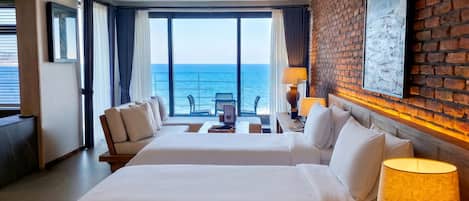 A hotel room with two beds, a balcony with a view of the sea, a brick accent wall, and a framed picture.