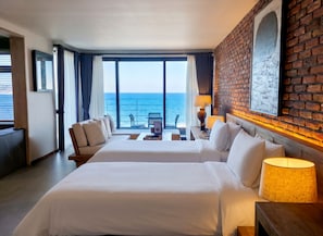 Habitación de hotel con dos camas, un balcón con vista al mar, una pared de detalles en ladrillo y un cuadro enmarcado.