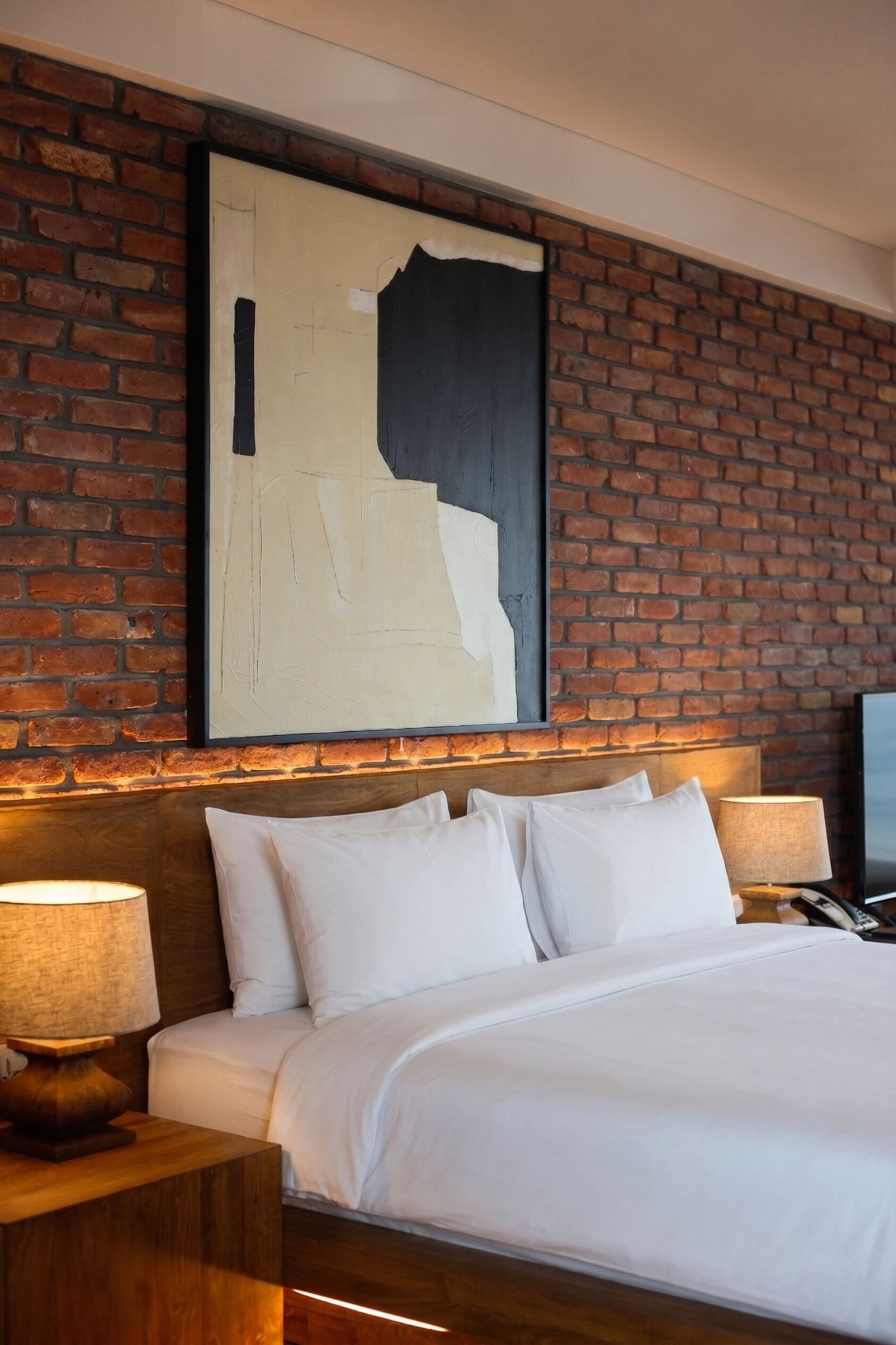 A hotel room with a brick wall, a large abstract painting, a wooden headboard, a bed with white linens, and two bedside lamps.