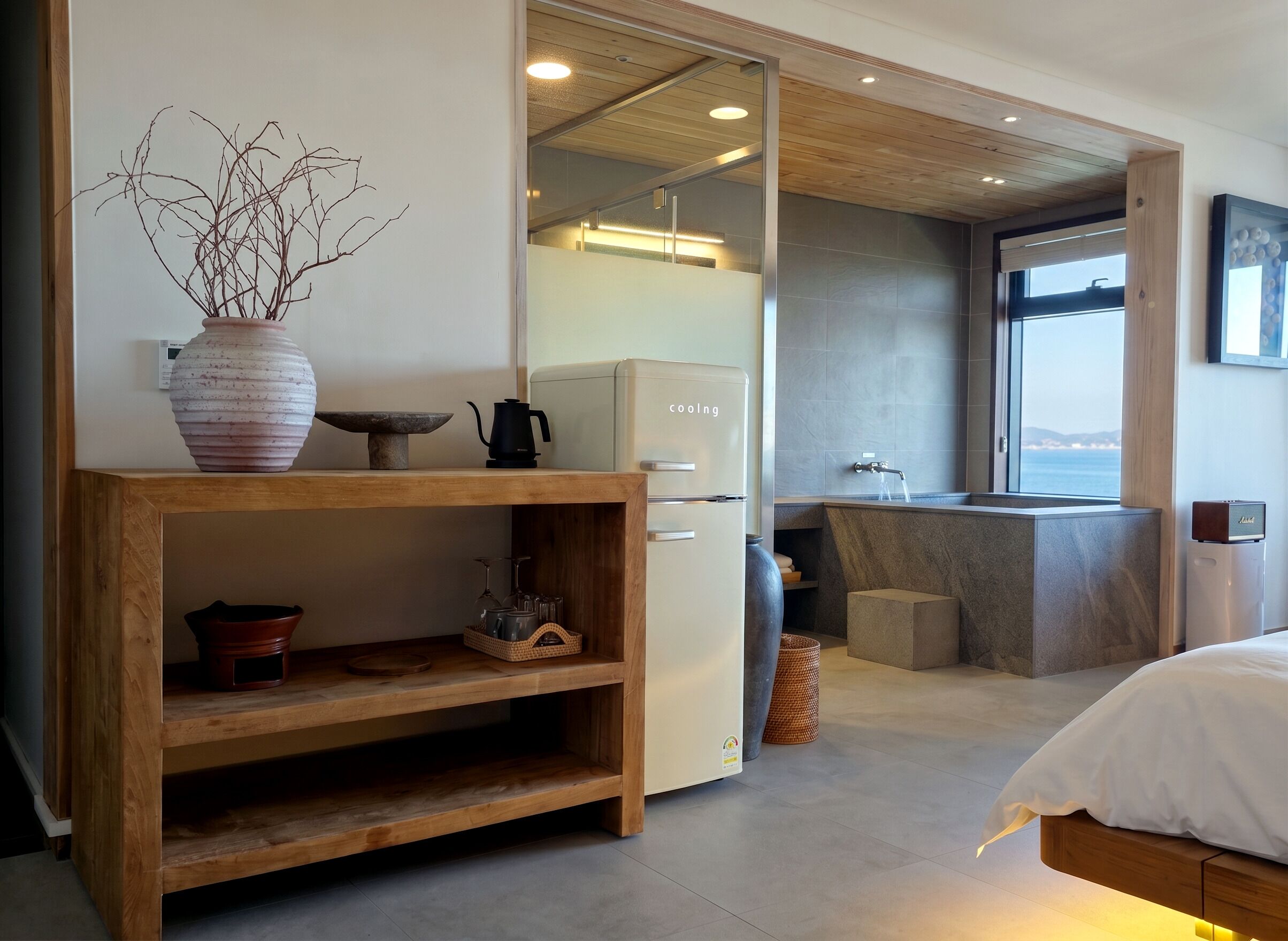 A modern hotel room with a wooden console table, a bed, a kitchen area with a sink, and a view of the sea.