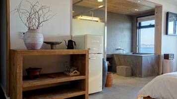 A modern hotel room with a wooden console table, a bed, a kitchen area with a sink, and a view of the sea.