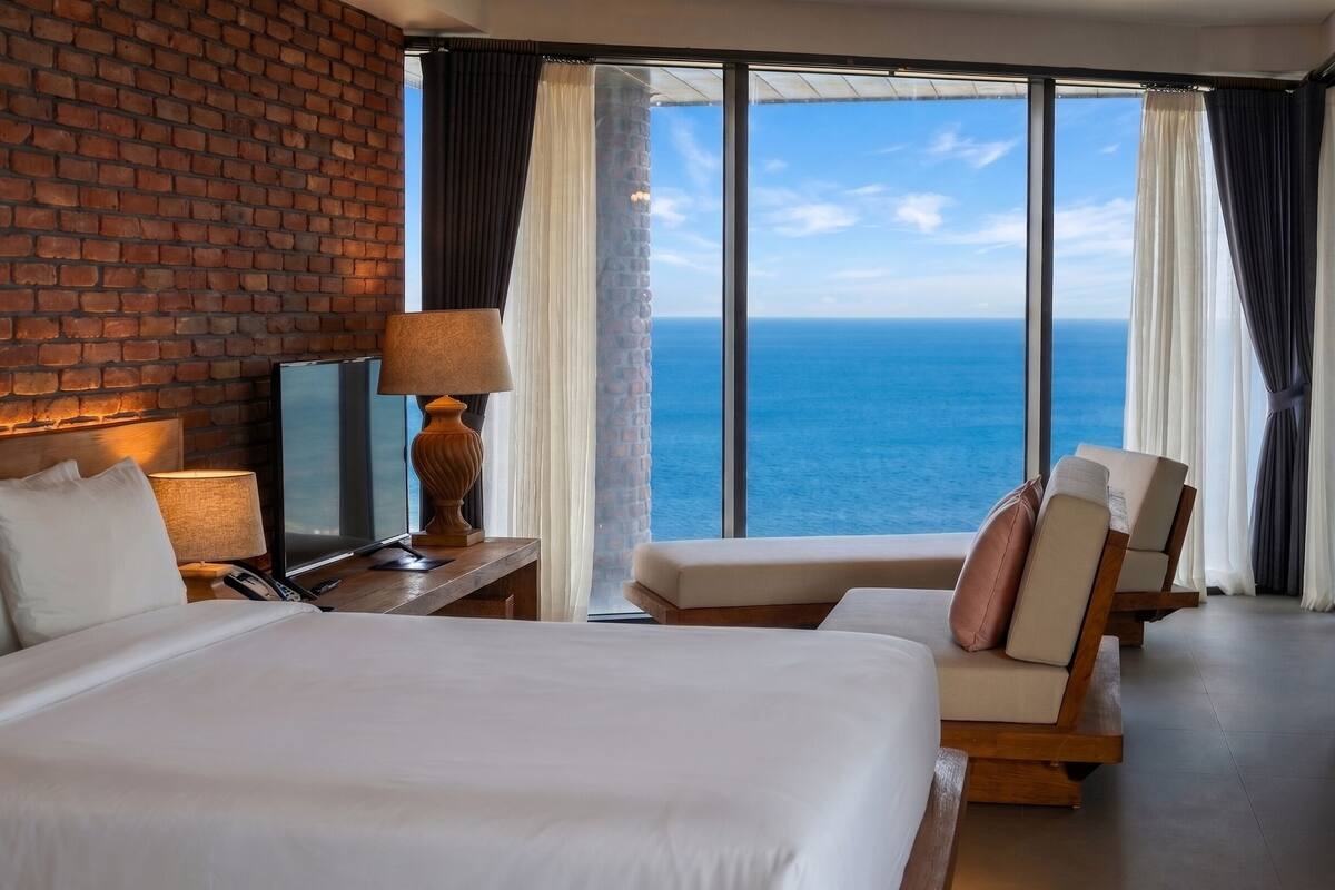 Une chambre d’hôtel avec un mur en briques, une grande fenêtre donnant sur l’océan, un lit, un bureau avec une lampe et une chaise.