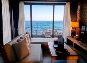 Habitación con sofá, mesa, lámpara y televisión. Se ve el océano a través de ventanas amplias.