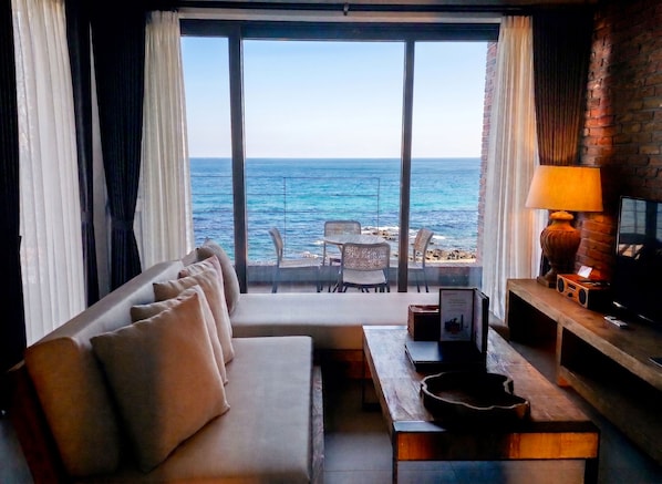 Habitación con sofá, mesa, lámpara y televisión. Se ve el océano a través de ventanas amplias.