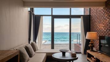 A modern living room with a sofa, a small round table, and a television. The room has large windows with curtains, offering a view of the sea.