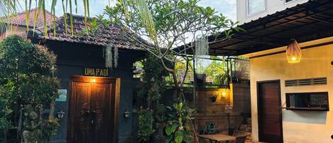 A traditional building with a wooden door labeled "UMA PADI," a small outdoor seating area, and a stone statue.