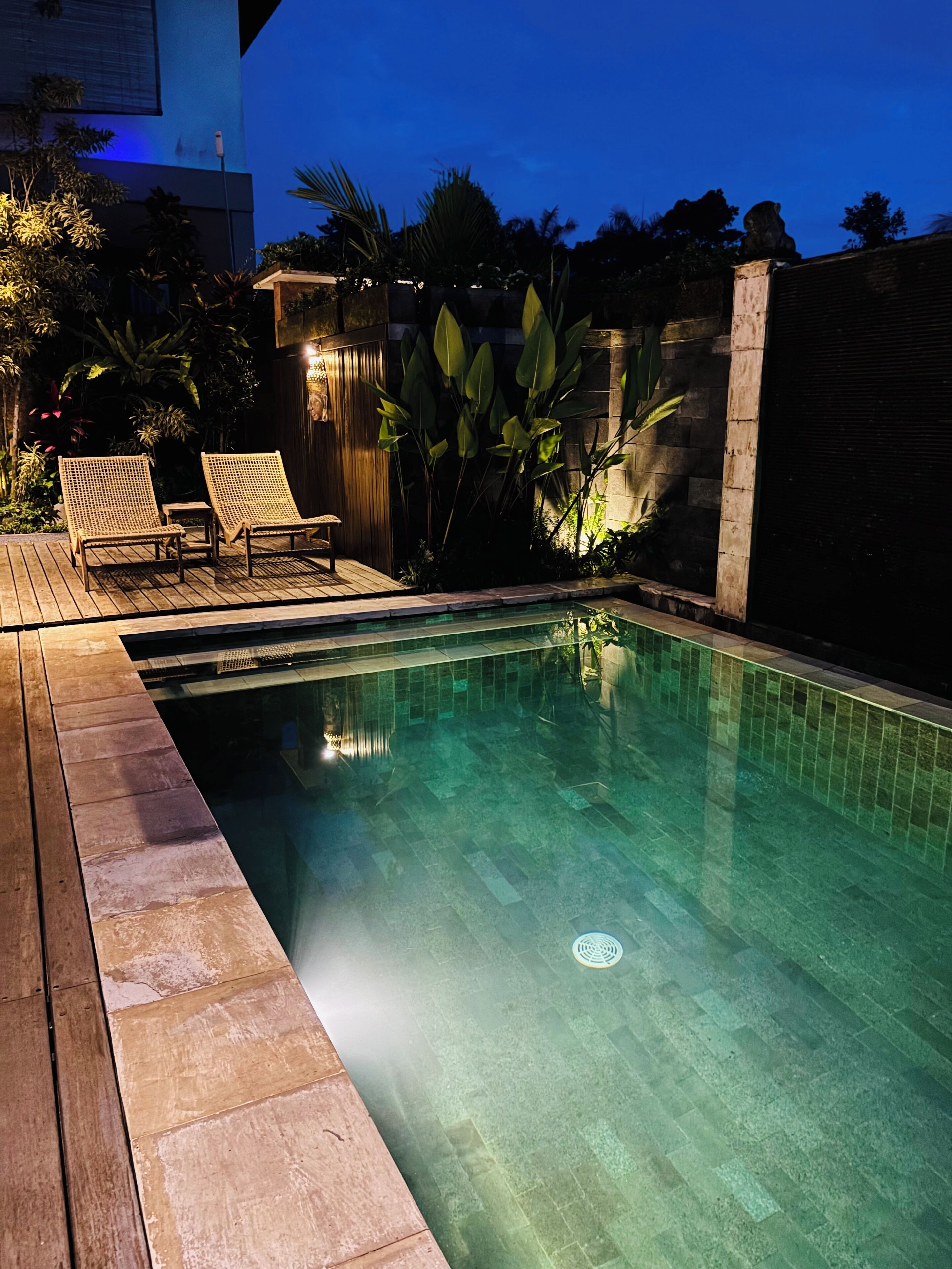 A clear swimming pool with a wooden deck and lounge chairs, surrounded by tropical plants and a stone wall.