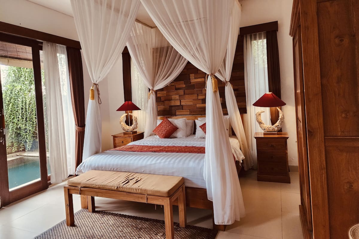 A bedroom with a four-poster bed, wooden headboard, two bedside tables with lamps, a bench, and a view of a swimming pool.