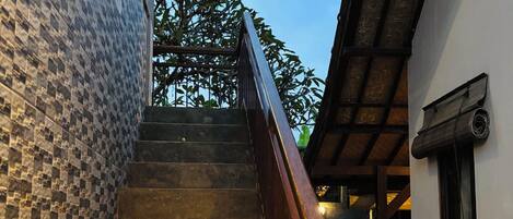 A staircase with a wooden handrail leading to a building with a tiled exterior wall and a window with a curtain.