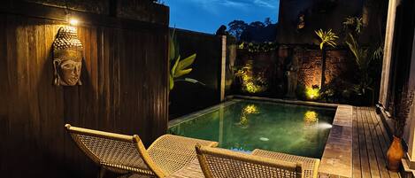 A wooden deck with a pool, two wicker chairs, and a Buddha head sculpture.