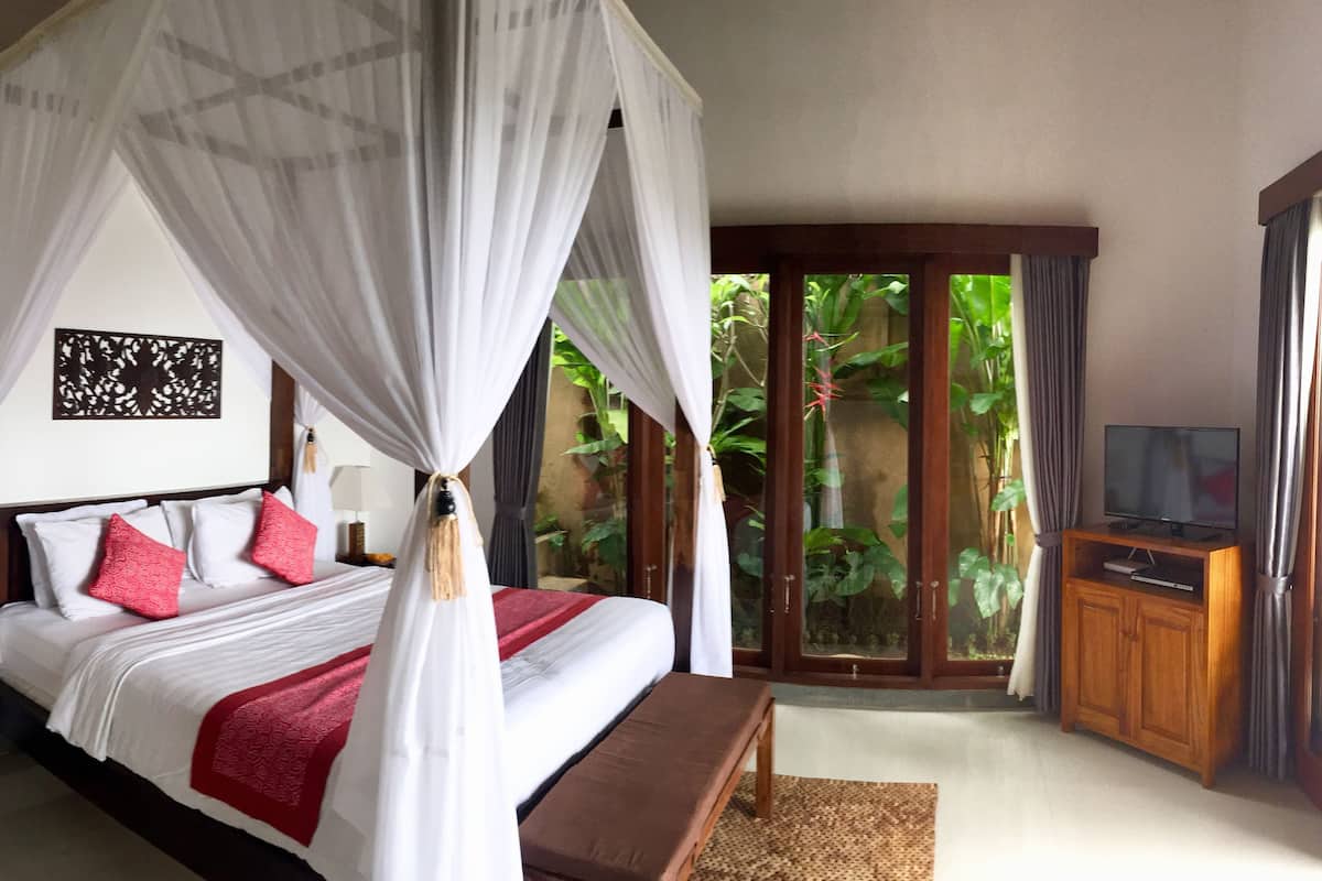 A bedroom with a four-poster bed, a wooden nightstand, a bedside table, a bench, a television on a wooden stand, and a view of greenery through a glass door.