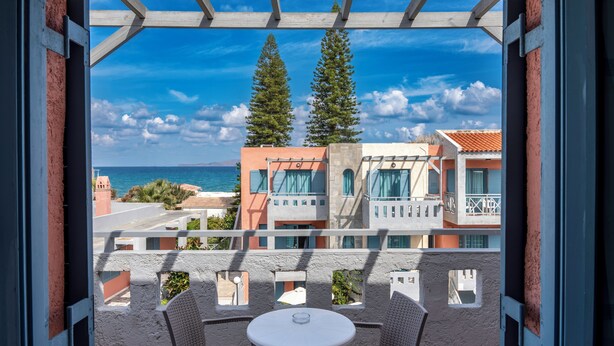 A balcony with a table and chairs, overlooking a coastal area with buildings and trees. - Marilisa Hotel (Hersonissos)