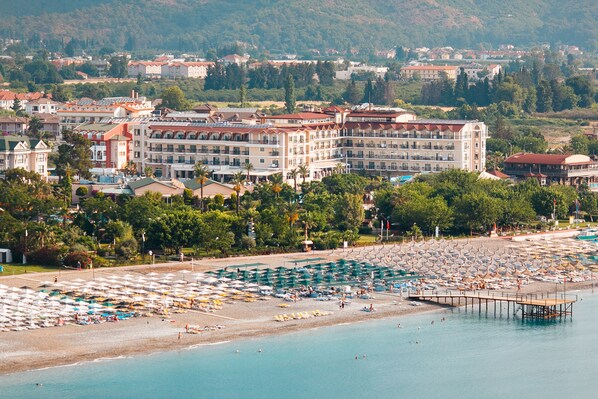 A coastal hotel with multiple floors, a beach with umbrellas, and a pier extending into the water. - L'Oceanica Beach Resort Hotel - All Inclusive (Kemer)