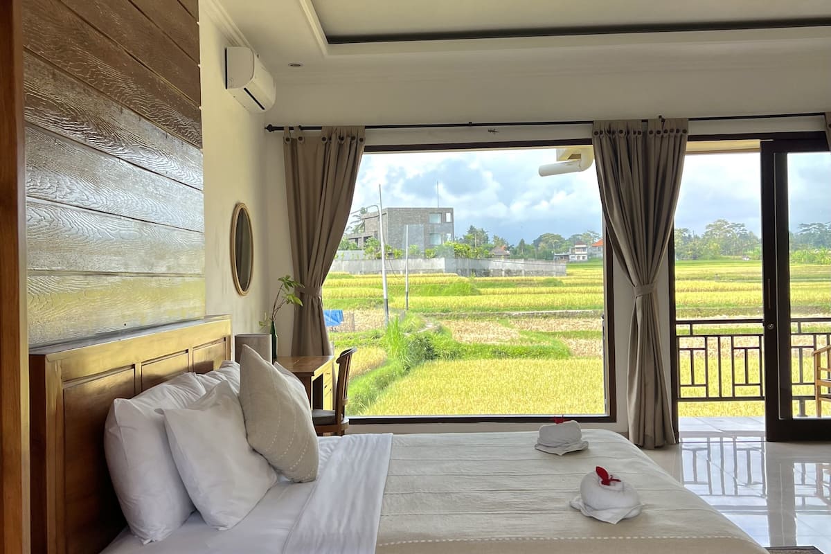 A bedroom with a large bed, wooden headboard, and a view of a rice field through the window.