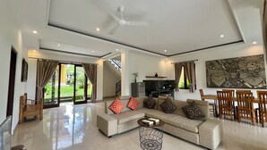 Amplia sala de estar con un sofá grande, una zona de comedor con sillas de madera, una cocina al fondo y un ventilador de techo.