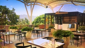 Un café al aire libre con mesas y sillas, rodeado de vegetación y un paisaje montañoso.