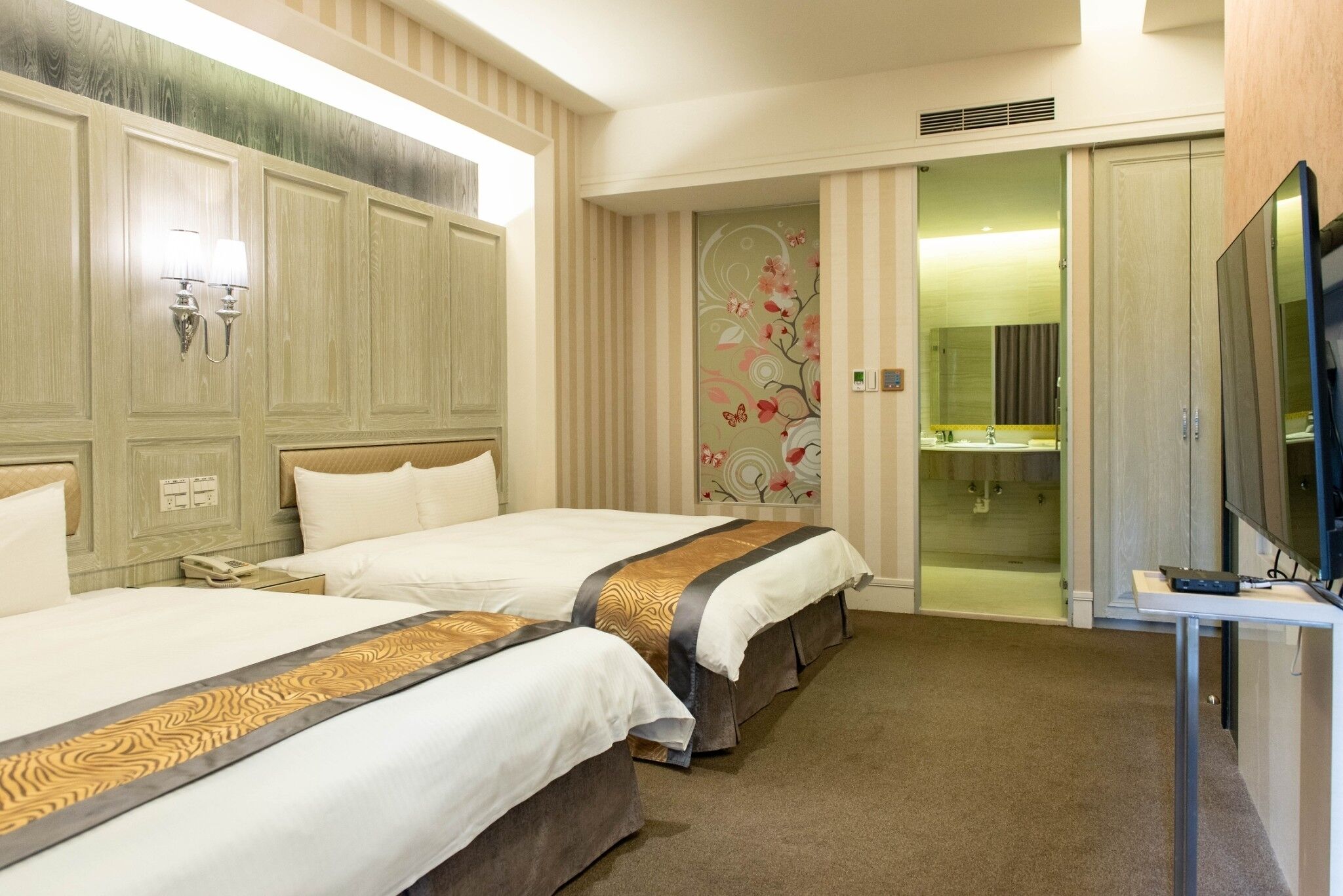 A hotel room with two beds, a television, a bathroom with a shower, and a mirror.