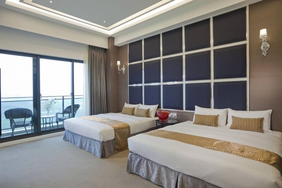 A hotel room with two beds, a large window with a view, and a balcony with chairs.