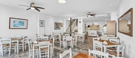 A spacious dining area with white chairs and wooden tables, ceiling fans, and a staircase leading to a kitchen area.