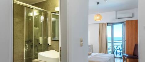A modern hotel room with a glass-enclosed shower, a large mirror, a wooden floor, a bed, a desk, and a view of the sea through a large window.