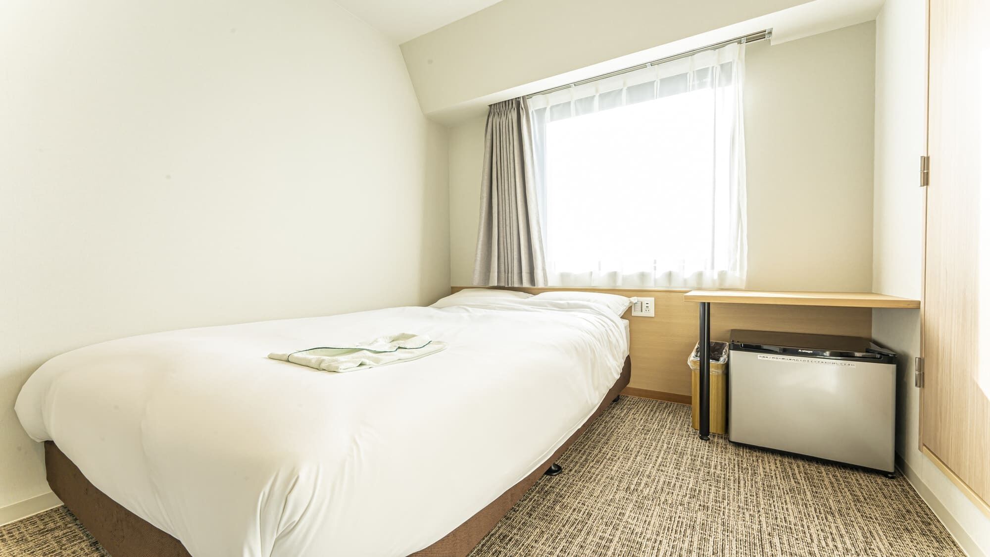 A hotel room with a single bed, a small desk, a trash bin, and a window with curtains.