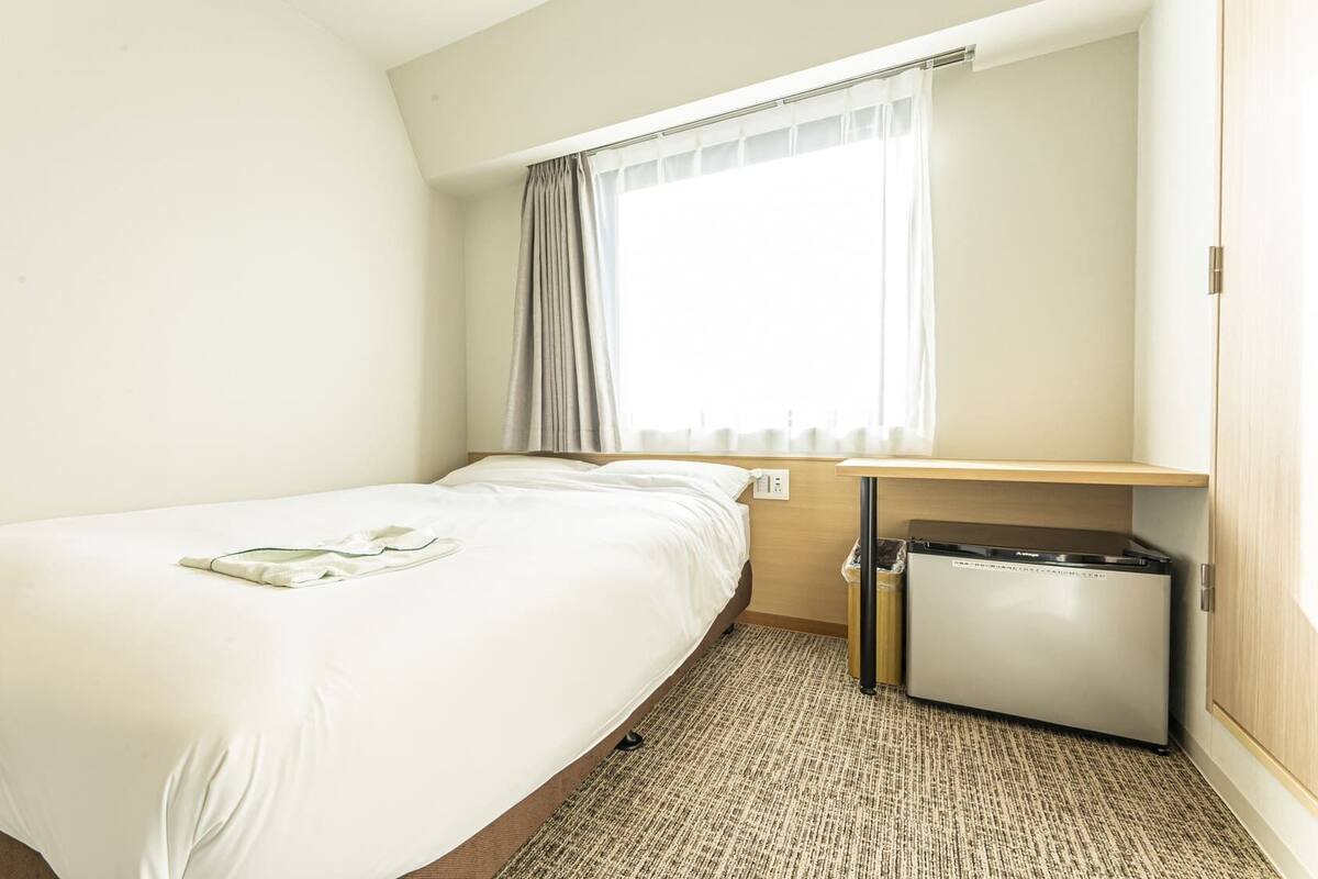 A hotel room with a single bed, a small desk, a trash bin, and a window with curtains.