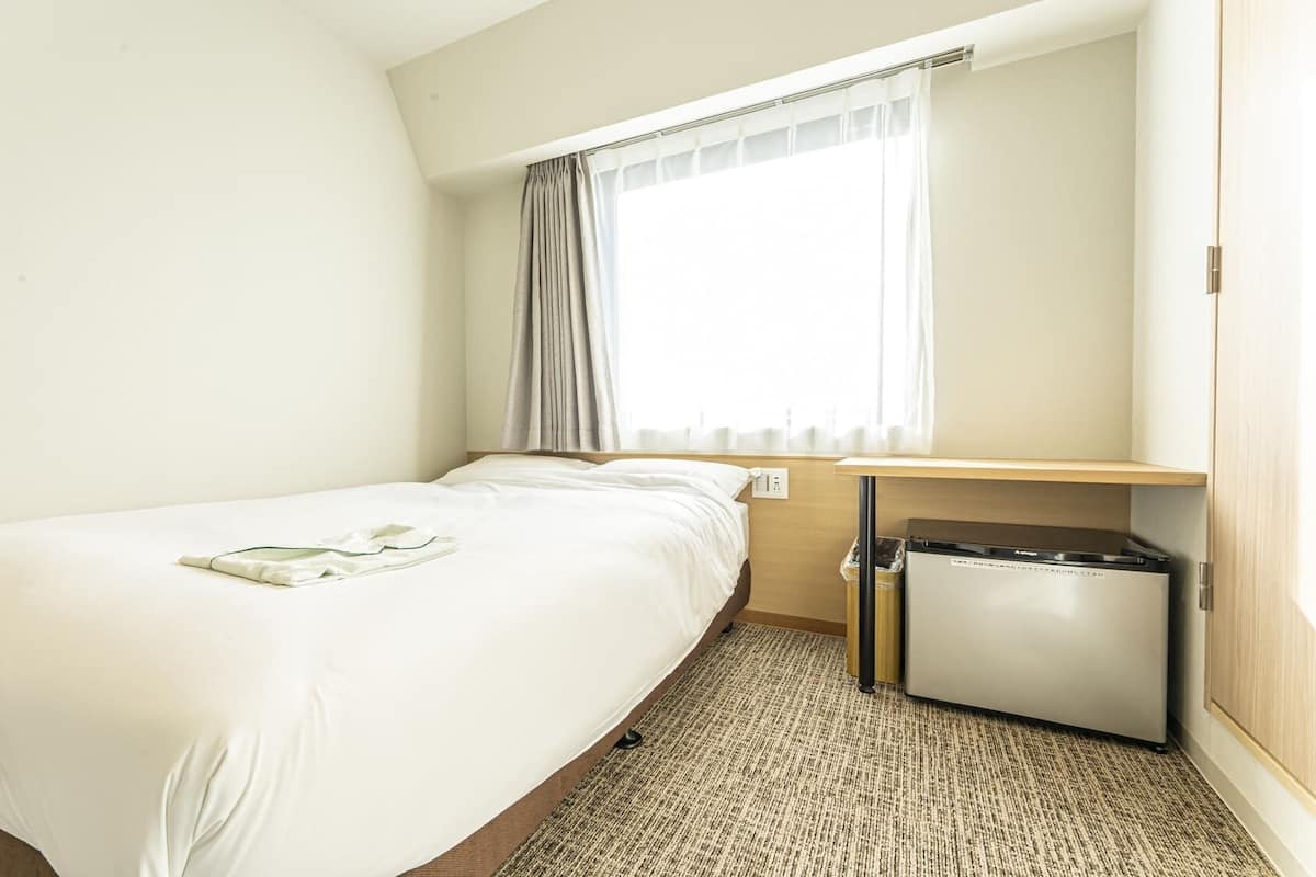 A hotel room with a single bed, a small desk, a trash bin, and a window with curtains.