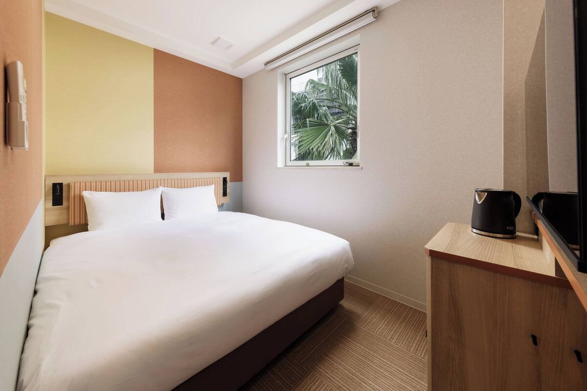 A hotel room with a bed, a wooden nightstand, a window with a view of palm trees, and a wall-mounted thermostat.