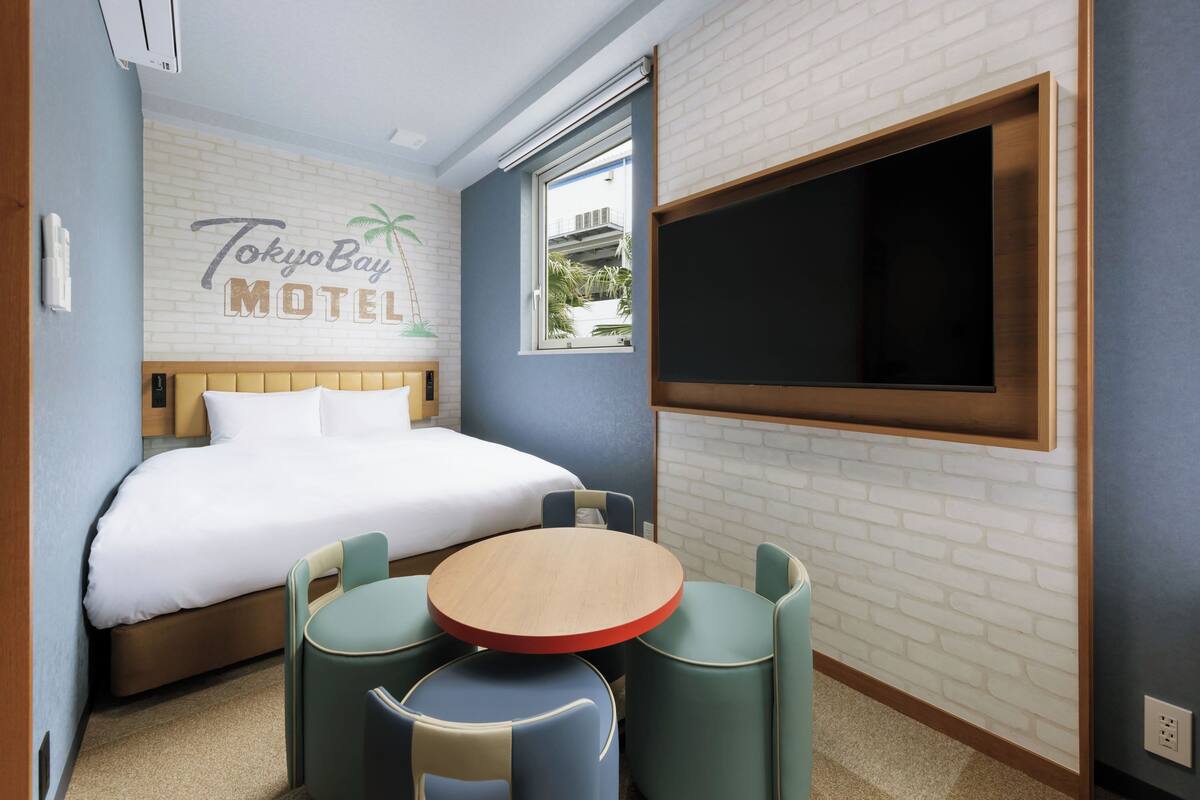 A hotel room with a bed, a round table with chairs, a TV mounted on the wall, and a brick-patterned wall with a palm tree and hotel name.