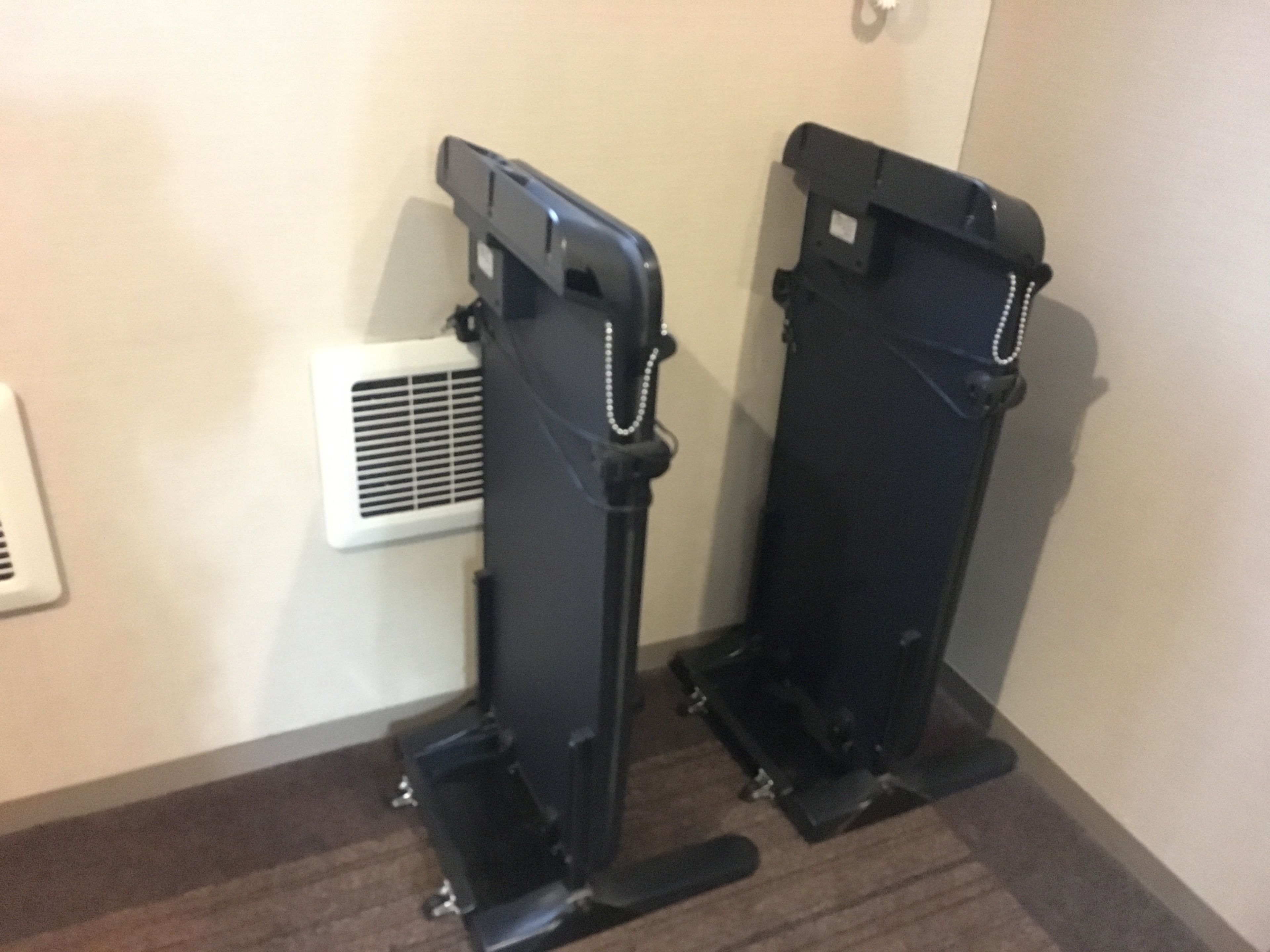 Two exercise machines in a room with a white vent and a brown floor.