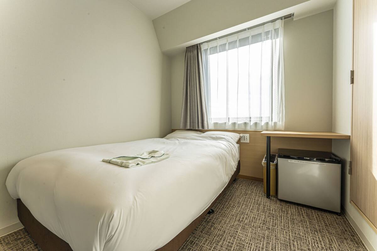 A hotel room with a single bed, a small desk, a compact refrigerator, and a window with curtains.