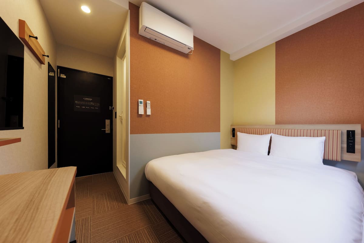 A hotel room with a bed, a desk, an air conditioning unit, a door, and a wall-mounted control panel.