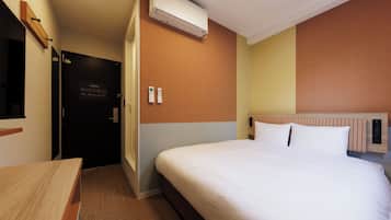 A hotel room with a bed, a desk, an air conditioning unit, a door, and a wall-mounted control panel.