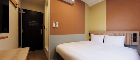 A hotel room with a bed, a desk, an air conditioning unit, a door, and a wall-mounted control panel.