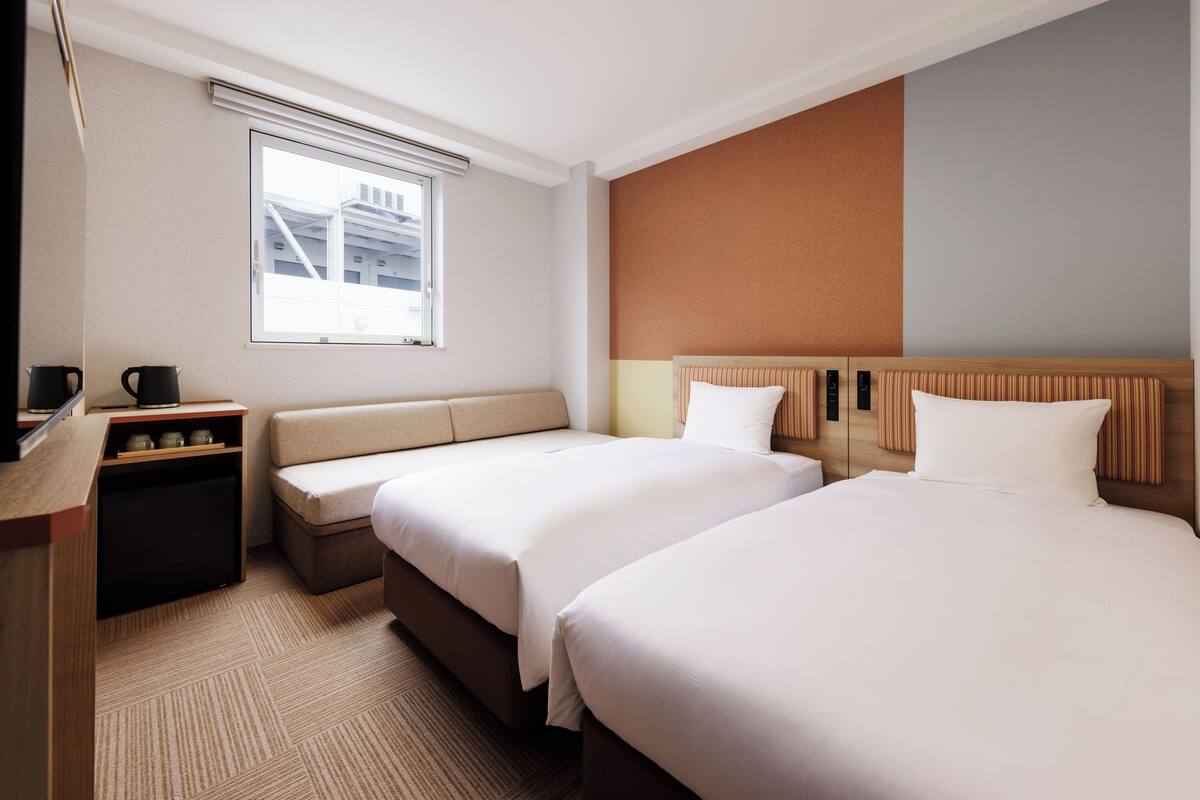 A hotel room with two beds, a sofa, a desk with a coffee maker, and a window with a view of buildings.