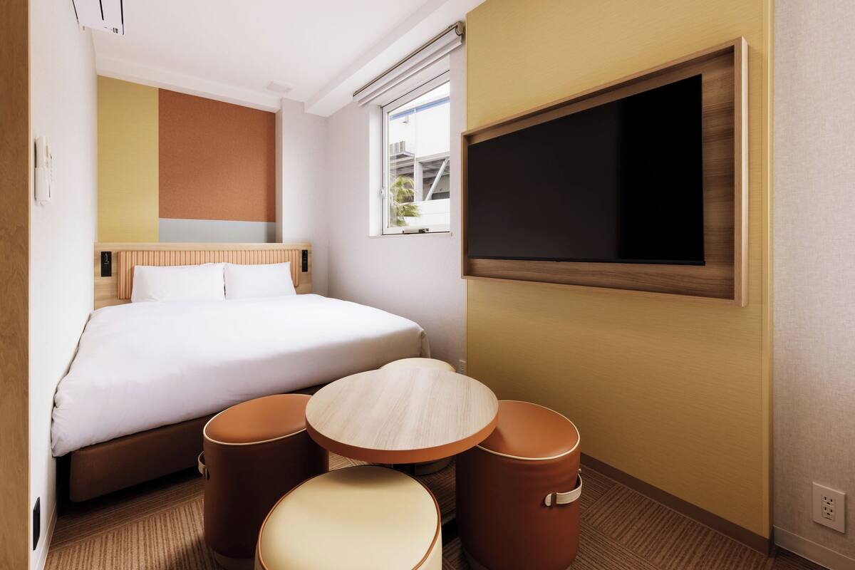 A hotel room with a bed, a small table, two stools, a TV, and a window with a view.