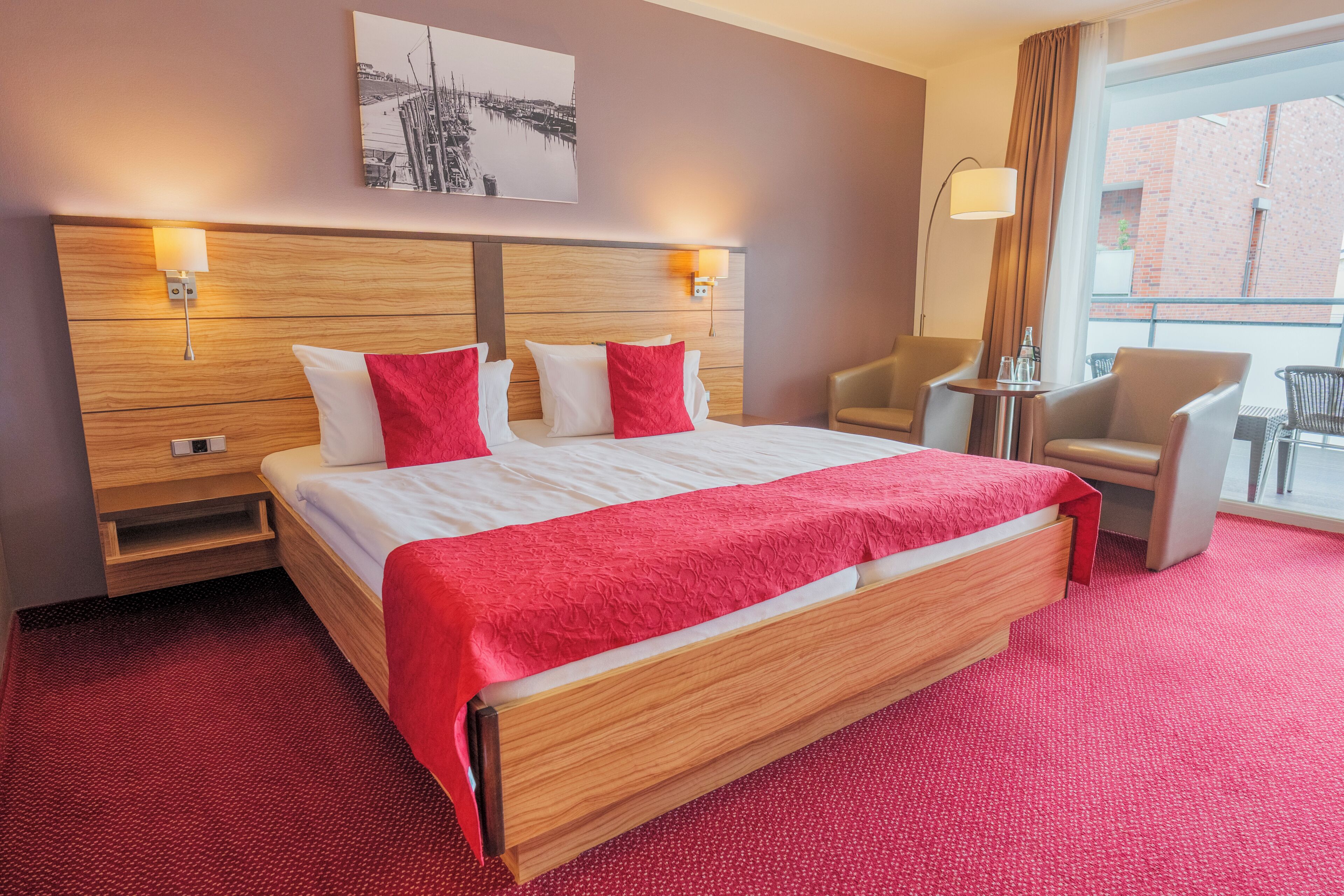Habitación de hotel con una cama grande, dos mesitas de noche, una silla, una pequeña mesa y una ventana con vista a un edificio.