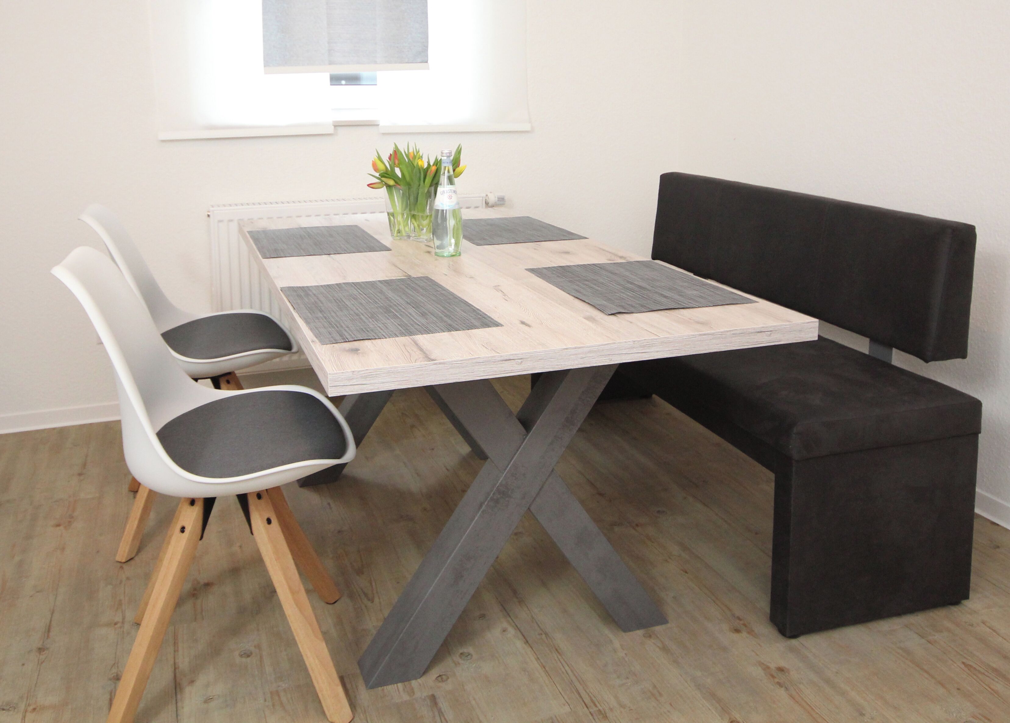 Una mesa de comedor con tapa de madera y patas metálicas, flanqueada por dos sillas blancas con cojines grises y un banco negro.