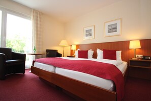 A hotel room with a large bed, two bedside lamps, a wooden headboard, a chair, a small table, and two framed pictures on the wall.