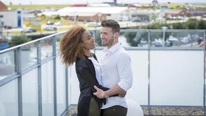 Una pareja se abraza en un balcón con vista a un puerto deportivo con barcos y edificios al fondo.