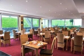 A restaurant with multiple tables and chairs, large windows with a view of greenery, and a red carpeted floor.