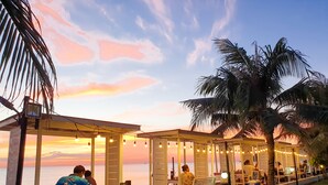 A beachside restaurant with outdoor seating, palm trees, and a sunset view. - Saint Tropez Beach Resort Hotel (Tha Mai)