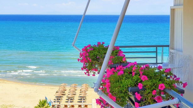 A balcony with a view of the ocean, a beach with umbrellas, and pink flowers. - Panorama Suites & Spa (Agia)