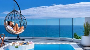 A person relaxing in a hanging chair by a pool with a view of the ocean. - Panorama Suites & Spa (Agia)