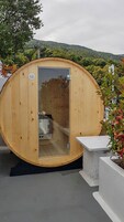 Una sauna de madera con puerta de vidrio, un banco en el exterior y una vista a una colina boscosa al fondo.