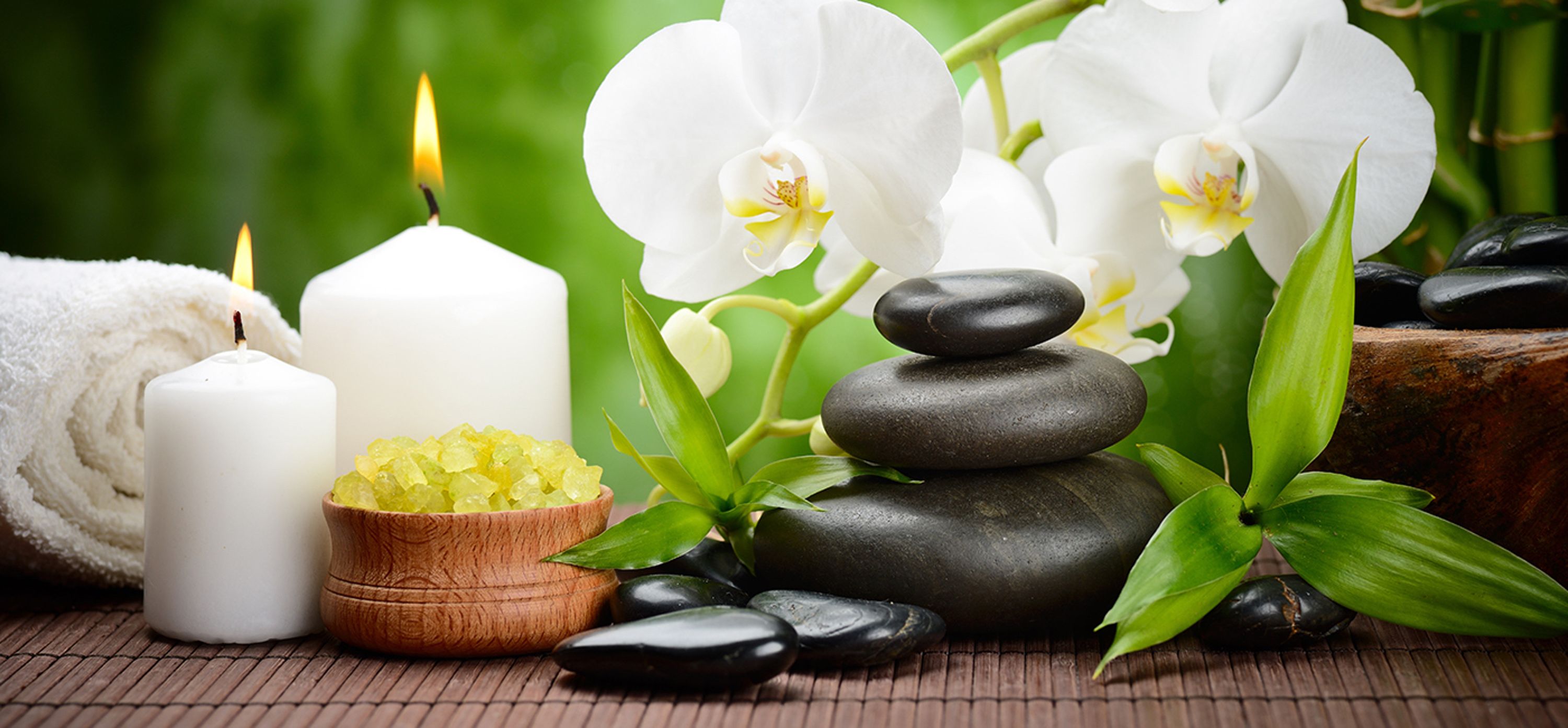 Un ambiente de spa con velas, orquídeas blancas, un cuenco de madera con sales de baño y piedras apiladas.