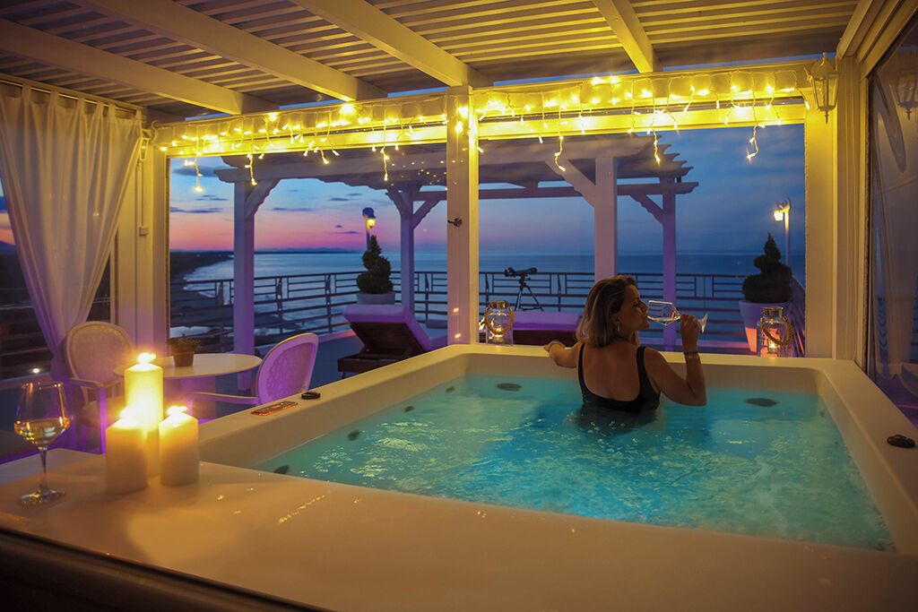 Una persona en un jacuzzi con velas y luces colgantes, con vistas a un atardecer.