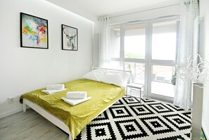 A modern bedroom with a bed, two framed artworks, a window with curtains, and a patterned rug. - LIVIN Apartments (Szczecin)